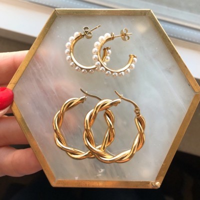 Dois pares de brincos tipo argola, um torcido dourado e outro dourado com pérolas, sobre apoio hexagonal branco e dourado.