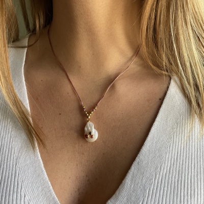 Colar com pendente branco irregular e bolinhas vermelhas e douradas num cordão castanho, usado por mulher com blusa branca decote V.