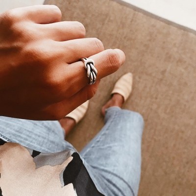 Mão com anel prateado trançado e roupa casual em fundo com tapete bege