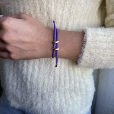 Pulseira roxa com fecho dourado no pulso de uma pessoa com camisola branca cremosa e calças azuis