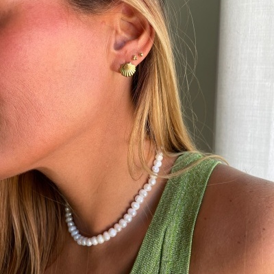 Brincos dourados com concha usados por mulher com colar de pérolas e vestido verde