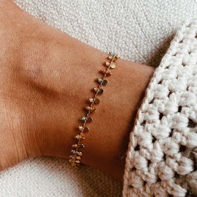 Pulseira dourada com pequenos elementos em formato de folhas no pulso de uma pessoa ao lado de roupa de crochê