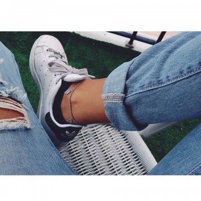 Pessoa com jeans rasgados e ténis Adidas brancos sentada numa cadeira branca sobre relva artificial