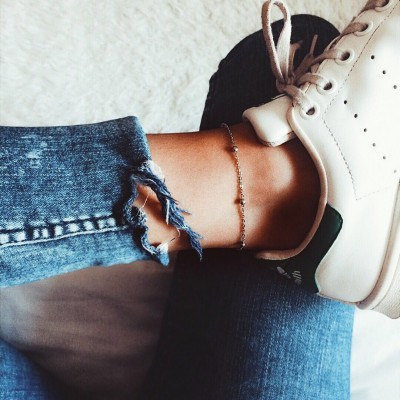 Perna com calças de ganga azul rasgadas, sapatilhas brancas e pulseira de tornozelo prateada