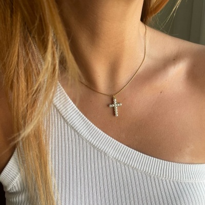 Colar dourado com pendente de cruz com pérolas usado por pessoa com blusa branca de tecido canelado