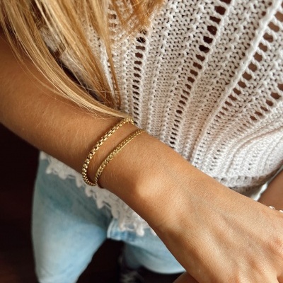 Pulseira dupla dourada em corrente fina no pulso de pessoa com blusa branca de malha, jeans e ténis pretos