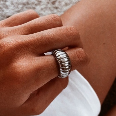 Mão feminina com anel prateado largo sobre roupa branca e fundo de tecido