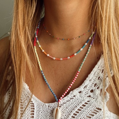 Três colares coloridos com contas, um com concha, em mulher com blusa branca de croché