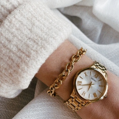 Relógio Seiko dourado com bracelete metálica, pulseira dourada, usado num pulso com camisola branca.