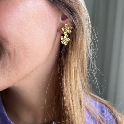 Brincos dourados em forma de flor numa mulher com cabelo loiro e camisola roxa