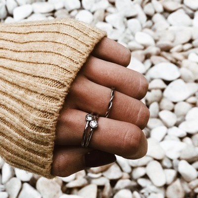 Mão com anéis prateados e camisola bege sobre pedras brancas.