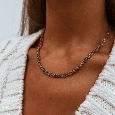 Colar em malha dourada trançada no pescoço de uma pessoa com suéter branco de malha.