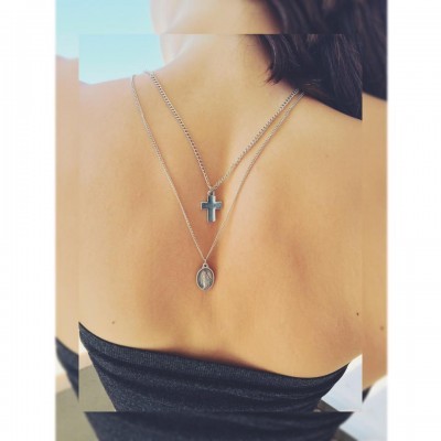 Colar duplo com cruz preta e medalhão oval em pescoço de pessoa com pele clara e cabelo castanho.