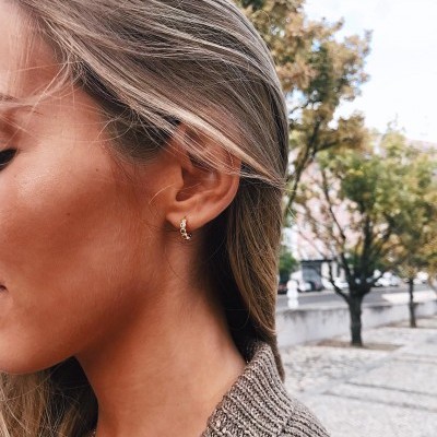 Brincos pequenos dourados em forma de argola com três bolas num ambiente urbano