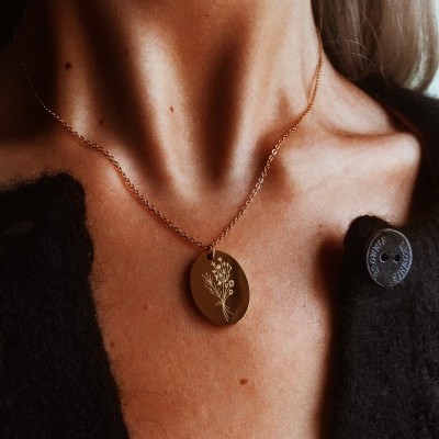 Colar dourado com pendente oval gravado com ramo floral no pescoço de pessoa com casaco preto