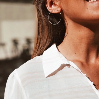 Brincos de argola prateados e camisa branca num fundo desfocado.