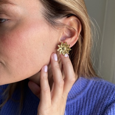 Brincos dourados em forma de flor na orelha de uma mulher loira com suéter azul.