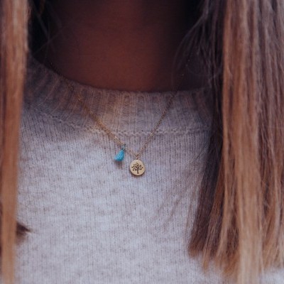 Colar dourado com pendentes azul e redondo usado por pessoa com suéter cinzento