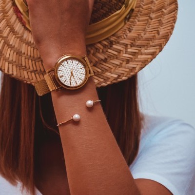 Relógio dourado com mostrador branco e pulseira dourada com pérolas, pessoa protegendo o rosto com chapéu de palha