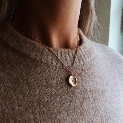 Colar dourado com pendente redondo usado por pessoa com camisola castanha felpuda