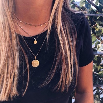 Pessoa com cabelo loiro usando t-shirt preta e três colares dourados com pendentes circulares