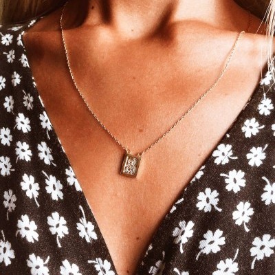 Colar dourado com pendente retangular com letras gravadas e vestido preto com flores brancas