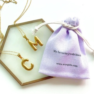 Colares com letras douradas C e M em base hexagonal e saco lilás com texto