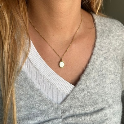 Colar dourado com pendente oval pequeno em pescoço de mulher com camisa branca e suéter cinza