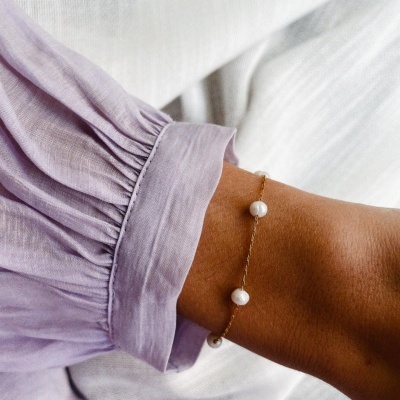 Pulseira dourada com pérolas no pulso de pessoa com blusa lilás