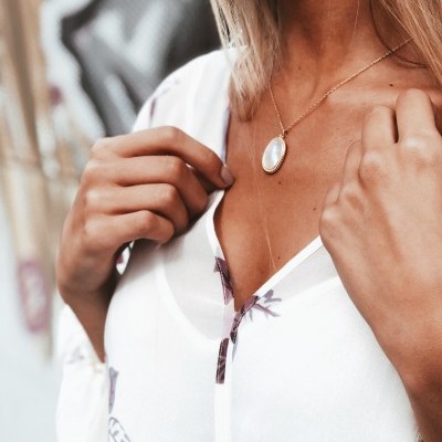 Colar oval branco com corrente dourada, usado com blusa branca com flores e parede grafitada ao fundo.