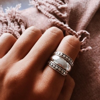 Anel prateado com bolinhas em dedo sobre tecido rosa claro com franjas.