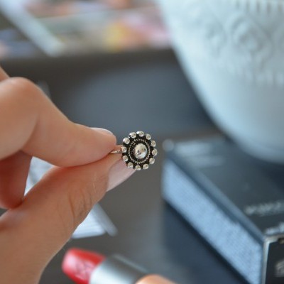 Anel circular prateado com pedras e pérola central, segurado entre dedos, com caixa preta e batom vermelho ao fundo