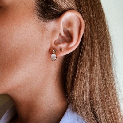 Orelha feminina com três brincos dourados pequenos e uma pedra branca oval.