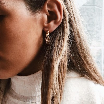 Brinco dourado de argola entrelaçada numa orelha de mulher com cabelo loiro