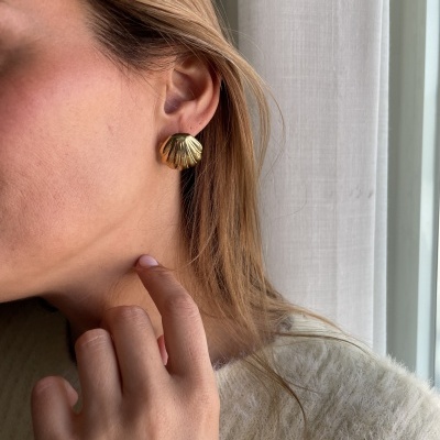 Brinco dourado em forma de concha usado por mulher perto de cortina branca