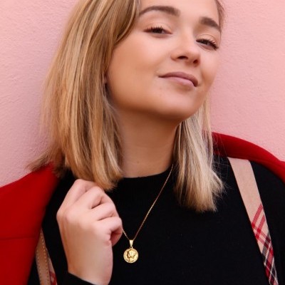 Mulher com casaco vermelho e colar dourado com pendente junto a parede rosa