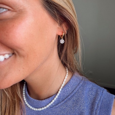 Brincos dourados com pendente flor branca e colar de pérolas em mulher com top azul.
