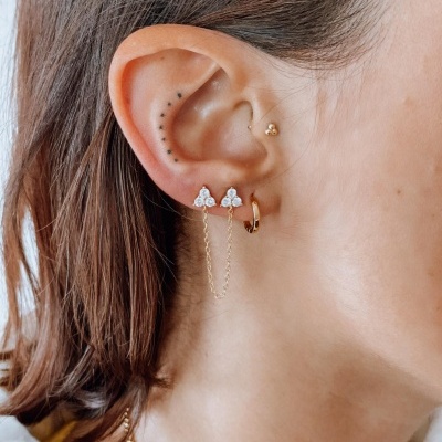 Orelha feminina com três brincos dourados, incluindo brinco com pedras claras e correntes finas douradas.