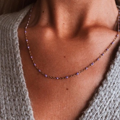 Colar fino dourado com contas lilás no pescoço de mulher com camisola cinza