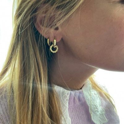 Brincos dourados em forma de coração pendurados em argolas, usados por mulher com cabelo loiro e camisola lilás com gola branca