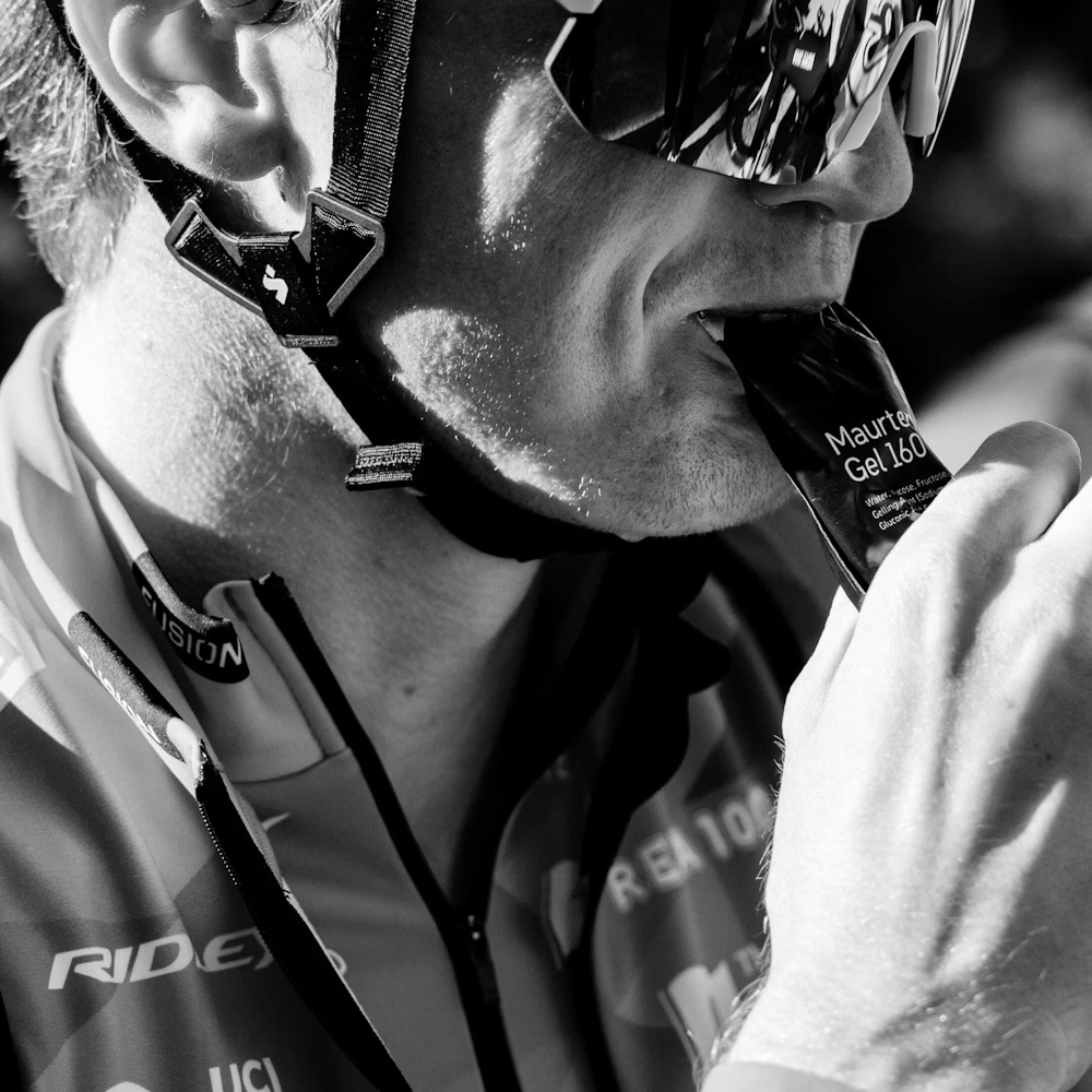 Ciclista a morder sachê de gel energético Maurten Gel 160 com capacete, óculos e roupa de ciclismo.