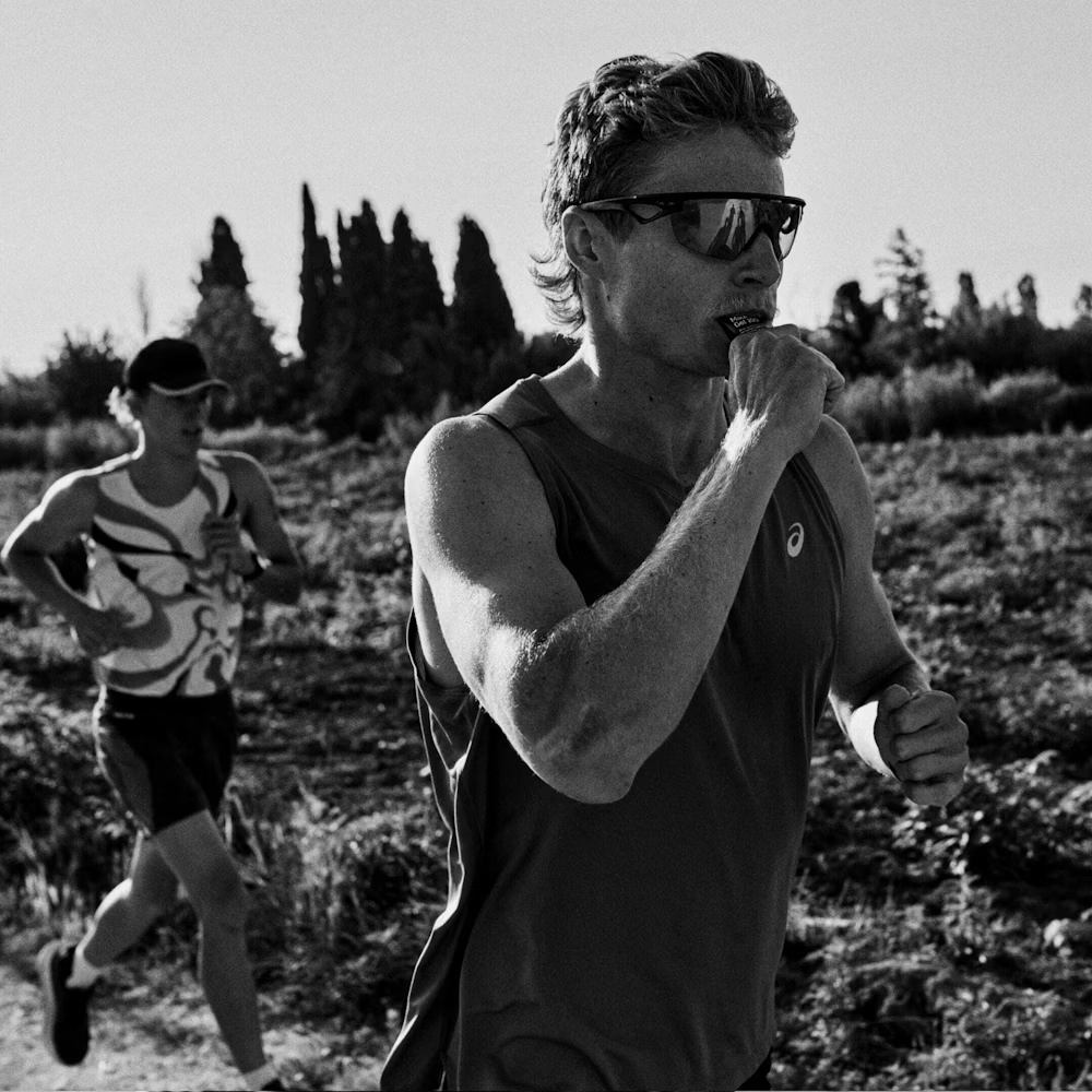 Homens a correr em exterior, um com óculos e camisola ASICS, a morder embalagem pequena