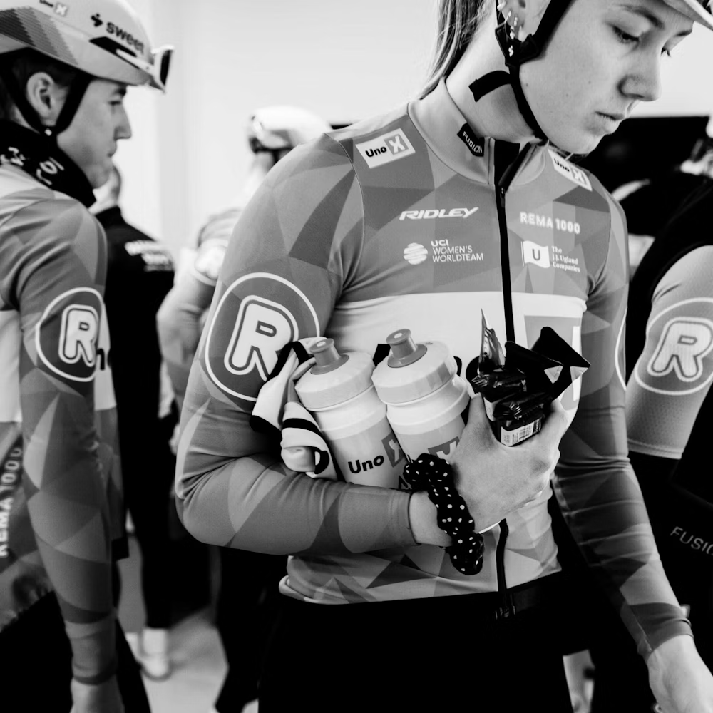 Atletas de ciclismo com fatos e capacetes, segurando garrafas e barras energéticas.