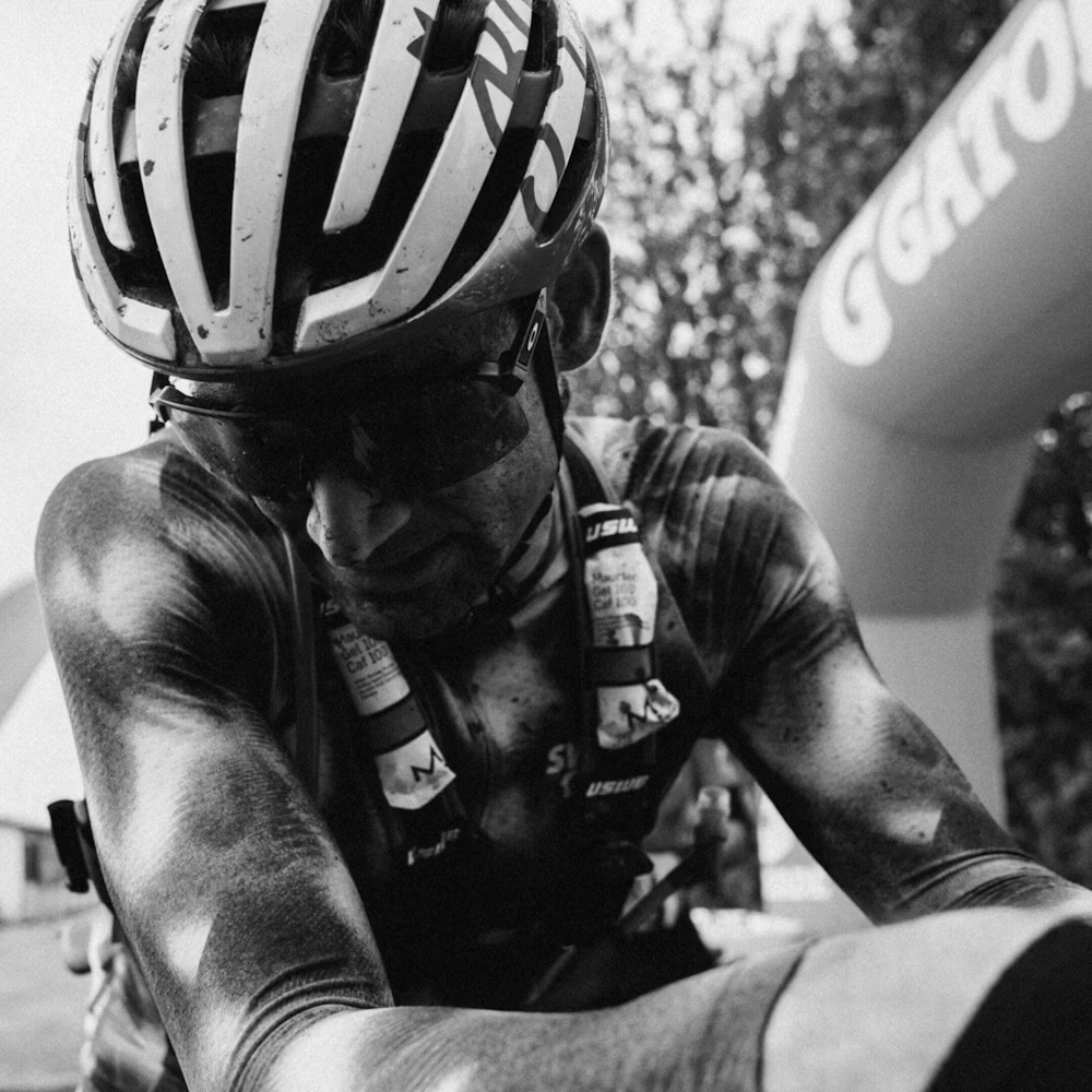 Ciclista com capacete branco e preto, óculos escuros e colete com gel energético num evento ao ar livre