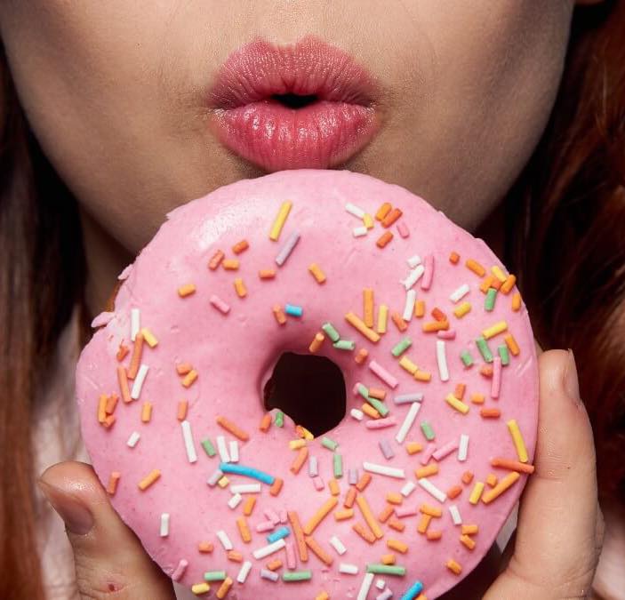 Pessoa segurando donut rosa com granulado colorido próximo aos lábios