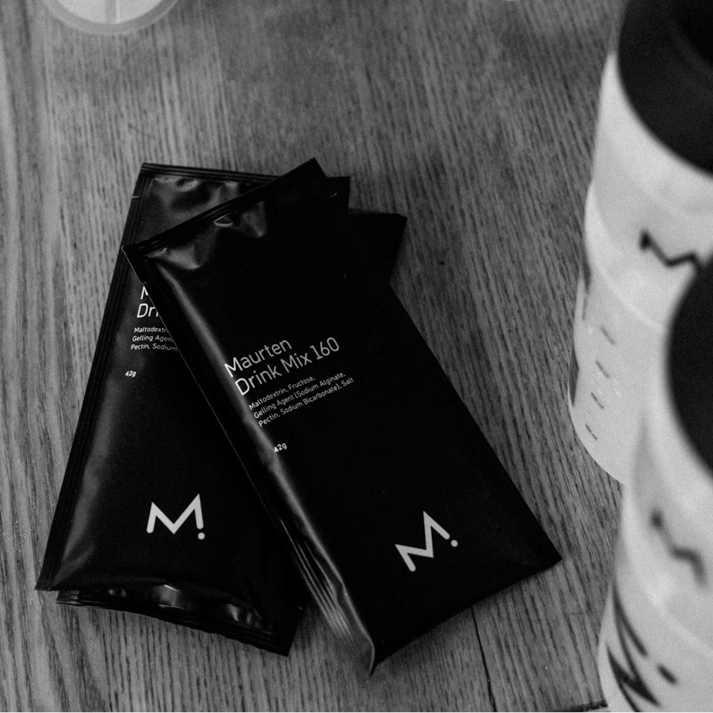 Sachets pretos de Maurten Drink Mix 160 em superfície de madeira com garrafas ao lado