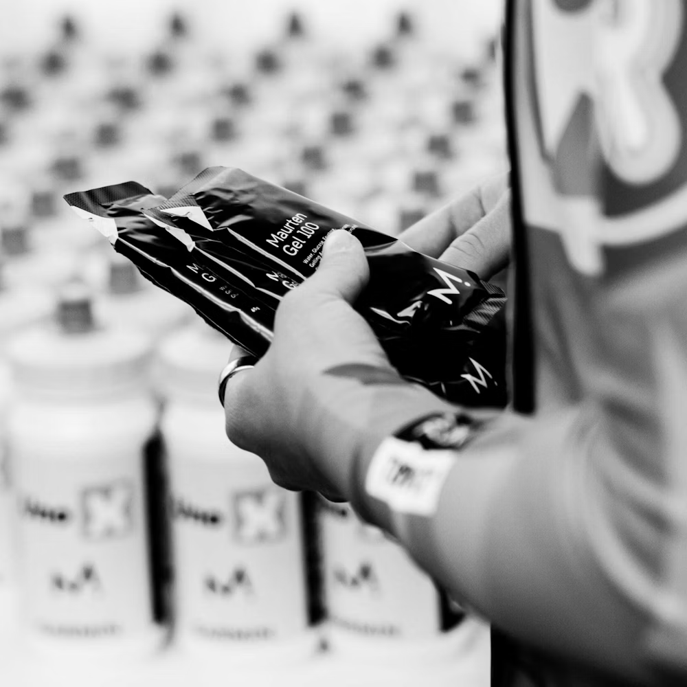 Pessoa a segurar pacotes pretos Maurten Gel 100 com garrafas brancas ao fundo