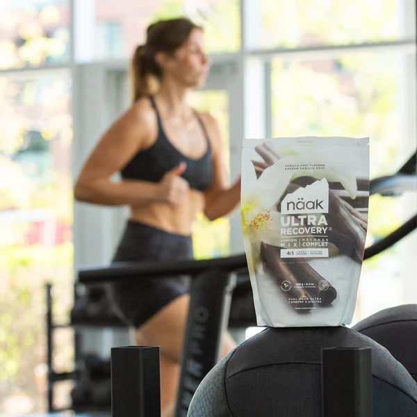 Pacote de suplemento nutricional branco 'naak ULTRA RECOVERY' sobre bola de exercício cinzenta num ginásio