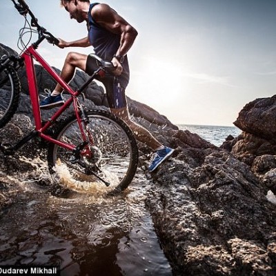 Homem com bicicleta de montanha vermelha em terreno rochoso perto do mar