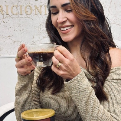 Mulher com casaco bege segurando chávena de café e frasco de suplemento na mesa